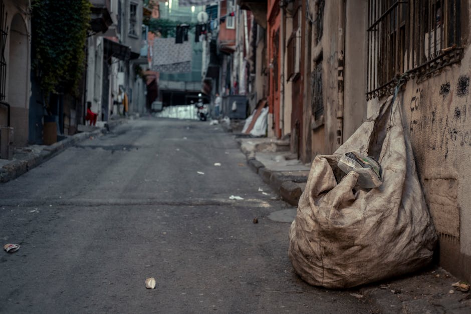 A large, worn-looking beige or light brown sack made of heavy-duty, weathered fabric is positioned on the right side of a narrow, uneven urban street. The sack appears filled with miscellaneous rubbish, with some loose debris, possibly plastic or paper, protruding from the top opening. The street surface is dark, asphalt with visible wear, cracks, and scattered small pieces of litter, including crushed drink cans and paper waste, which contribute to an overall appearance of untidiness. To the right of the sack, there is a weathered concrete or brick wall with blackened graffiti or dirt marks, and an iron fence with decorative bars mounted on it, partially obscured by the sack. In the background, along the street, there are closely packed residential buildings with a mix of concrete and brick facades, some with laundry hanging on clotheslines. The ambient lighting suggests a cloudy or overcast day, lending a slightly dull and subdued tone to the scene. The setting exemplifies a typical urban alley that might require private waste collection or rubbish removal services, such as those offered by Rubbish Removal Ealing, to maintain cleanliness and local environment standards.