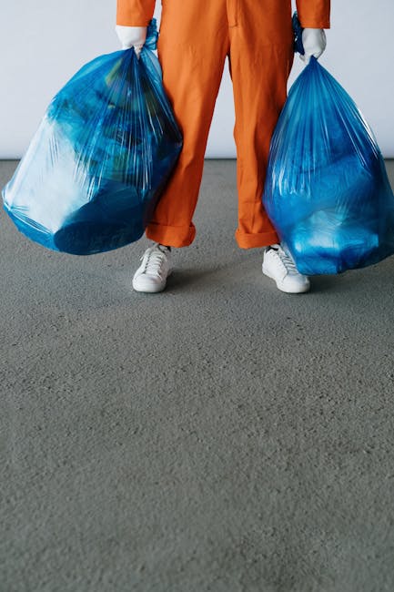 A person wearing orange coveralls and white sneakers is standing on a grey, textured surface indoors, holding two large, semi-transparent blue plastic rubbish bags filled with waste. The bags are slightly crinkled and bulging with household or general rubbish, with visible contents suggesting crumpled paper or lightweight debris. The background features a plain, light-colored wall, indicating an indoor environment such as a storage area or warehouse. The individual’s face and upper body are not visible, emphasizing the focus on the rubbish bags and their contents. This scene relates to waste collection, potentially representing private waste handling or alternative rubbish removal services operated by companies like Rubbish Removal Ealing, highlighting the typical equipment used in rubbish disposal and the physical process of rubbish collection at the ground level.