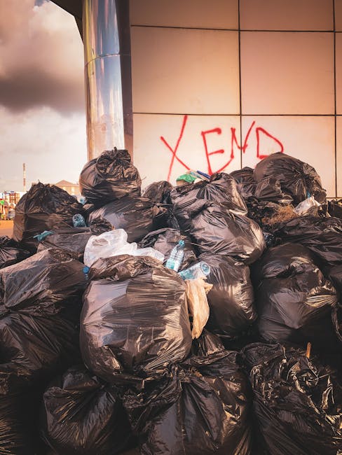 A large pile of black trash bags stacked against a building with a tiled wall, some of which are slightly torn or bulging, revealing glimpses of their contents. Several discarded plastic bottles, including clear water bottles, are visible among the bags. The bags occupy most of the foreground, with some leaning and overlapping each other, suggesting a recent waste collection or unattended rubbish. In the background, part of a metallic cylindrical structure, possibly a street lamp or utility pole, is visible on the left side, along with an overcast sky filled with gray clouds. The building's wall has a reddish-orange graffiti tag stating 'XEND' written in spray paint. The scene appears to be outdoors, likely on a pavement or street-side area near an urban commercial or residential building, indicating an instance of private waste disposal or rubbish accumulation outside a collection schedule managed by local authorities. The image reflects an environment requiring professional rubbish removal, such as those provided by companies like Rubbish Removal Ealing, to clear such waste effectively and safely in an urban setting.