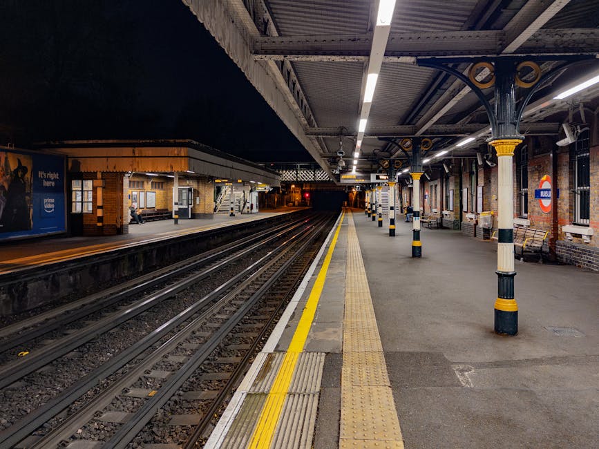 Underneath the elevated platform of Golders Green station, there is a luggage and passenger waiting area with a wooden bench in the foreground. Several individuals are seated on the benches, while others stand nearby, dressed in casual and winter attire. The platform is sheltered by a white canopy with a scalloped edge, supported by black metal beams. A black-and-yellow sign hanging from the canopy indicates the way out with an arrow, and a lift sign is also visible, suggesting accessible access points for passengers. In the background, part of the station’s infrastructure and rail tracks are visible, along with a large red and white roundel sign displaying the station name. The scene is illuminated by natural daylight, casting shadows on the ground, and the overall appearance suggests a typical urban railway station environment, consistent with a location that offers services related to transportation and potentially the collection or disposal of waste generated by travelers or station operations. Rubbish removal services like those provided by Rubbish Removal Ealing may handle waste clearance in such transportation hubs.