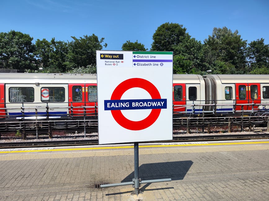 A white signboard with a large red circle logo and a blue horizontal bar displaying the text 'EALING BROADWAY' in white letters, positioned on a paved train station platform. The sign includes directional indicators for the way out, district line, Elizabeth line, and national rail and bus services, with small icons and text in black, blue, and red. Behind the sign, a modern train carriage with a silver body and red and blue stripe accents runs parallel to the platform, resting on railway tracks. Tall green trees with leafy canopies are visible further back, under a clear blue sky with bright sunlight casting shadows on the platform surface. The overall scene depicts an urban transportation hub, reflecting the infrastructure of public transit in London, with a focus on accessibility and station signage, relevant to independent and alternative methods of transport and waste management in busy locations.