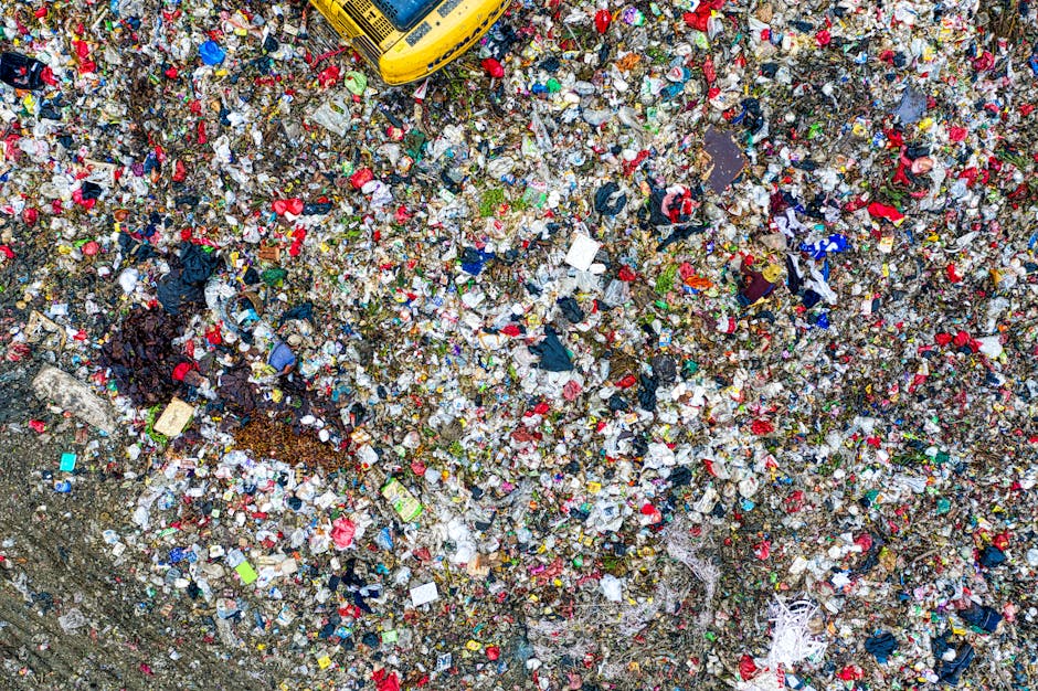 Aerial view of a large landfill site filled with a dense layer of mixed waste, including plastic bottles, paper, fabric, and other household refuse, creating a cluttered surface with various colors and textures. In the top right corner, part of a yellow waste collection vehicle is visible, indicating waste management activity. The waste appears compacted and uneven, with some areas showing slight mounds and patches of soil or debris beneath. The scene is outdoors, with natural daylight illuminating the heap, suggesting an environment where rubbish is either awaiting collection or being sorted for disposal. The overall setting symbolizes the scale of waste accumulation and highlights the importance of efficient rubbish removal services like those offered by Rubbish Removal Ealing, particularly in the context of alternative waste handling methods outside of regular household collection. This detailed depiction emphasizes the need for professional rubbish clearance in managing large quantities of waste responsibly.
